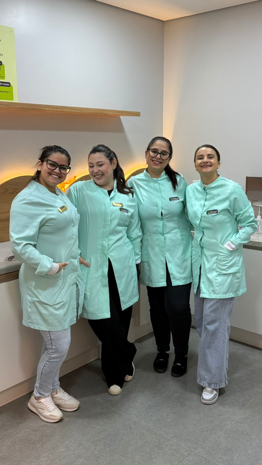 Quatro integrantes da equipe NH Vacinas de uniforme verde-água, sorrindo no interior da clínica