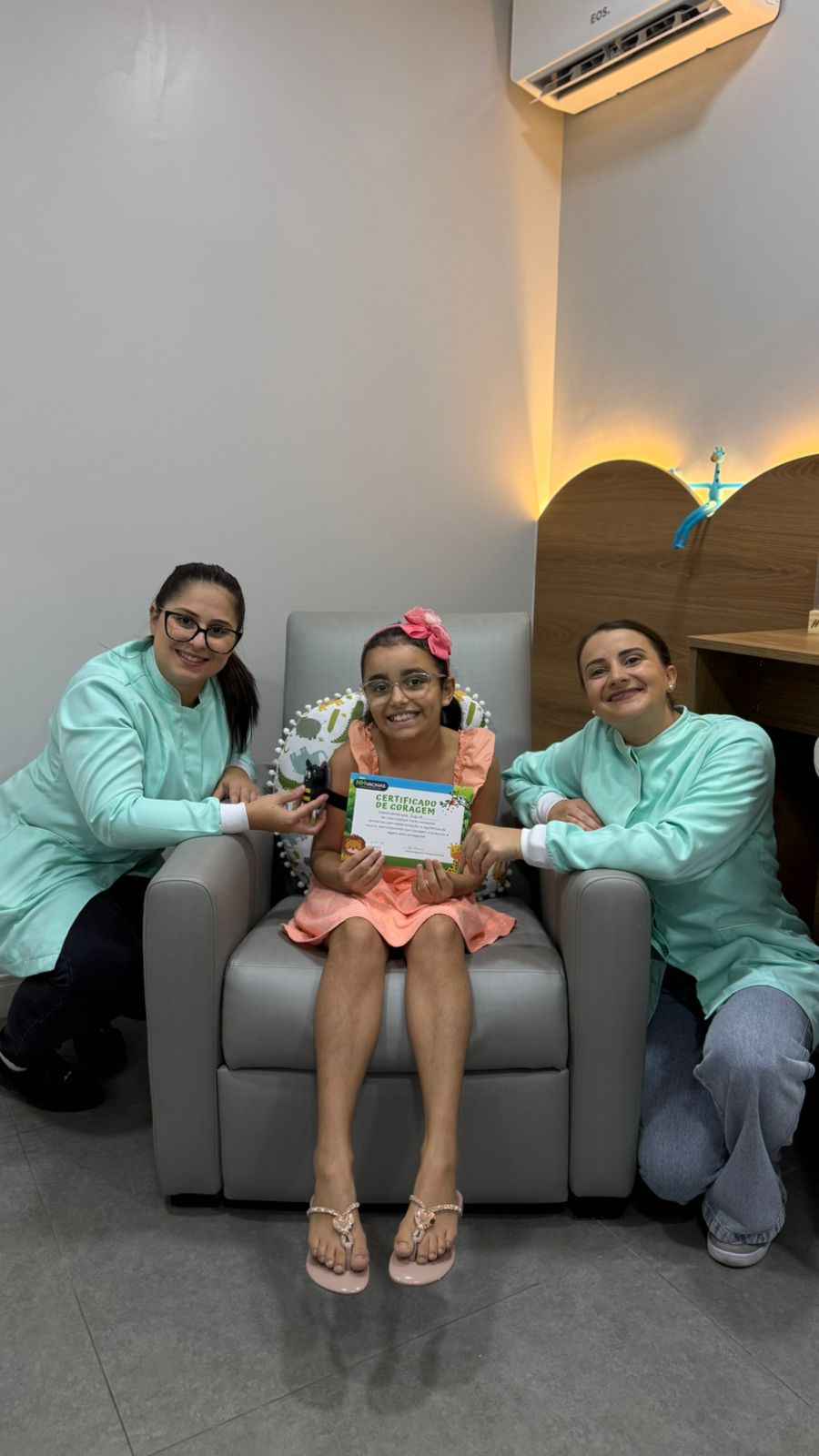 Menina sorridente sentada na poltrona da clínica NH Vacinas segurando o Certificado de Coragem, entre duas enfermeiras